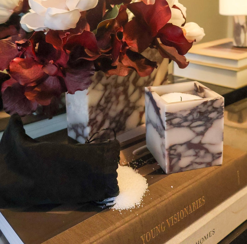 A square calacatta viola marble vessel next to a pouch filled with candle sand styled on books next to a marble vase