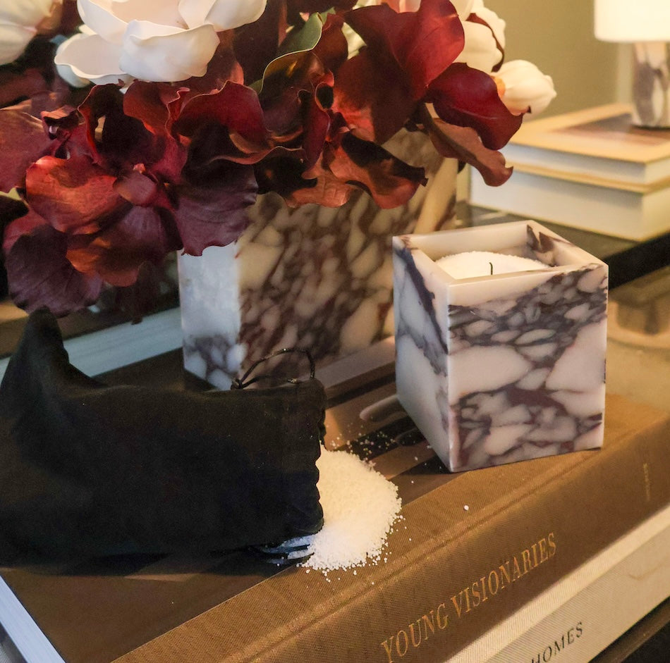A square calacatta viola marble vessel next to a pouch filled with candle sand styled on books next to a marble vase