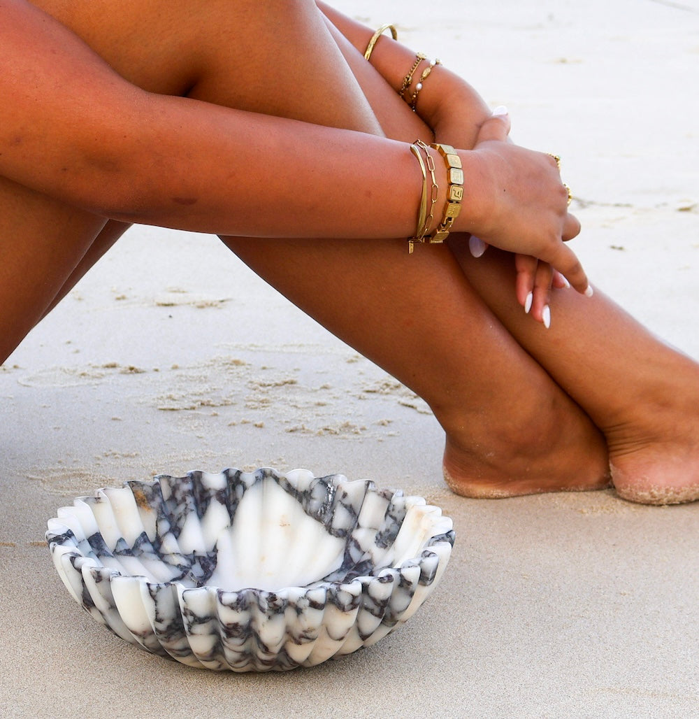 Sculptural Calacatta Viola marble bowl with soft scalloped edge on beach next to woman's legs